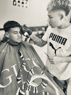 A person with tattoos and a Puma T-shirt is styling the hair of another individual who is wearing a barber cape with a snake design and the Gucci logo. They appear focused on a precision haircut. The room is equipped with chairs and a poster on the wall showing different hairstyles.