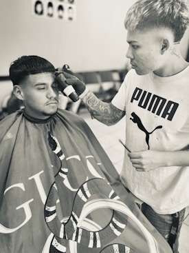 A person with tattoos and a Puma T-shirt is styling the hair of another individual who is wearing a barber cape with a snake design and the Gucci logo. They appear focused on a precision haircut. The room is equipped with chairs and a poster on the wall showing different hairstyles.