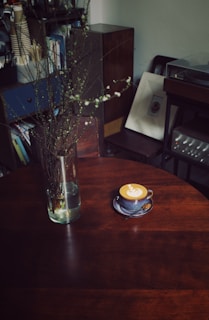 A cozy indoor setting with a wooden table holding a cup of latte art coffee on a saucer and spoon. Next to it is a glass vase containing branches with small white blossoms. The background includes books, a framed picture, and vintage audio equipment, suggesting a warm, lived-in space.