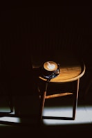 A wooden chair is placed in a dimly lit setting with a cappuccino on the seat. The coffee cup features intricate latte art on the foam, and a spotlight highlights the chair and cup, creating a dramatic contrast between light and shadow.