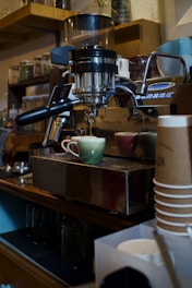 A coffee machine is preparing espresso in a cozy cafe setting. Two ceramic cups are positioned under the portafilter, one green and one red, as the coffee pours into them. Shelves in the background display an assortment of jars, likely filled with coffee beans or other ingredients. A stack of disposable coffee cups and other barista equipment such as pitchers and cups are visible nearby.