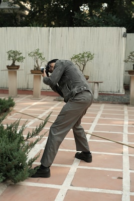 A person is leaning forward while taking a photograph in a garden setting. They are dressed in a black leather jacket and grey pants, framing their shot with a camera. The background consists of potted plants placed on stands against a wooden fence, creating a serene atmosphere. The foreground shows some green shrubs growing along the tiled pathway.