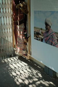 The image shows a section of a wall adorned with traditional textiles and garments. There is a photograph attached to the wall depicting a man in traditional attire, including a turban. The shadow patterns created by a lattice structure nearby add a decorative element to the floor.