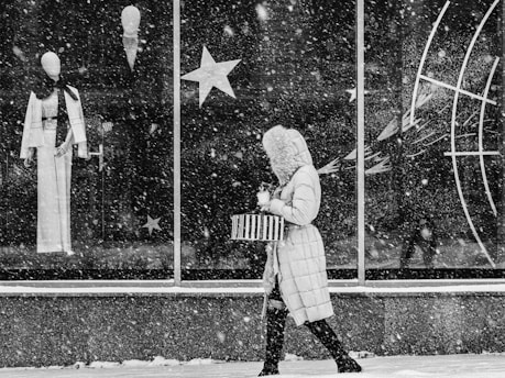 A person wearing a hooded winter coat carries a striped bag and a cup while walking past a large store window during heavy snowfall. The window displays mannequins dressed in stylish outfits, accompanied by star-shaped decorations.