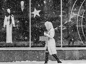 A person wearing a hooded winter coat carries a striped bag and a cup while walking past a large store window during heavy snowfall. The window displays mannequins dressed in stylish outfits, accompanied by star-shaped decorations.