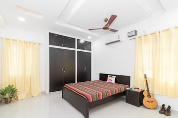 A modern bedroom with bright lighting and a minimalist design. The room features a double bed with a colorful striped bedsheet and a pillow with a mustache pattern. A dark wooden wardrobe with multiple compartments stands against one wall. Yellow curtains cover the windows, and a ceiling fan and air conditioning unit are installed. A guitar rests beside the bedside table, which holds a few books. A pair of shoes is placed on the floor near the guitar. A potted plant adds a touch of greenery to the room.