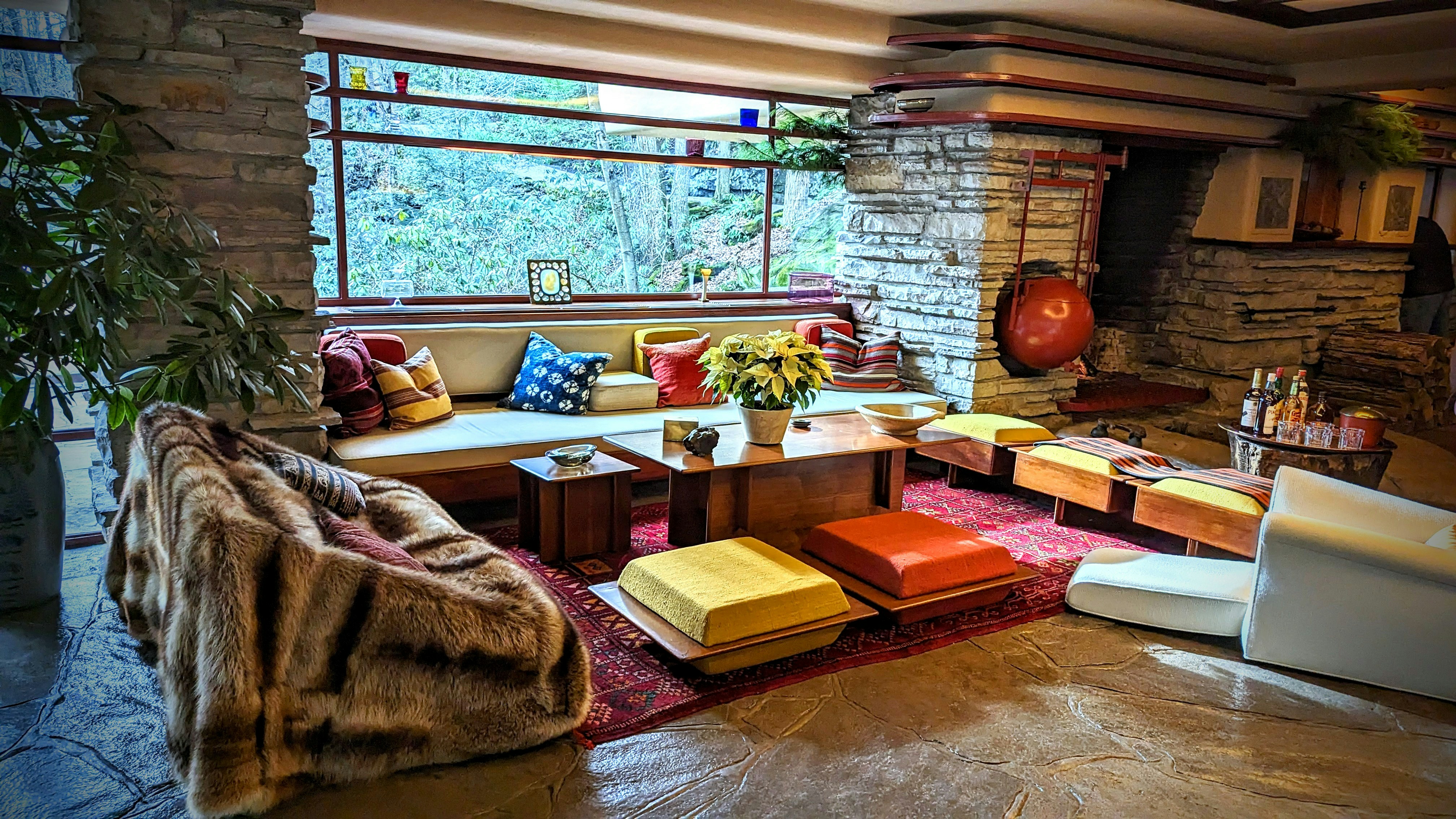 A stylish living room with a mid-century modern aesthetic features stone walls and a large, panoramic window that provides an expansive view of the natural surroundings. The space is furnished with a mix of colorful cushions, low seating, and a central wooden table adorned with a plant. Warm tones and textures, including a plush fur throw, complement the rustic stone fireplace in the background. The atmosphere is cozy and inviting, accentuated by strategically placed decorative items and soft lighting.