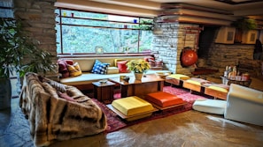 A stylish living room with a mid-century modern aesthetic features stone walls and a large, panoramic window that provides an expansive view of the natural surroundings. The space is furnished with a mix of colorful cushions, low seating, and a central wooden table adorned with a plant. Warm tones and textures, including a plush fur throw, complement the rustic stone fireplace in the background. The atmosphere is cozy and inviting, accentuated by strategically placed decorative items and soft lighting.