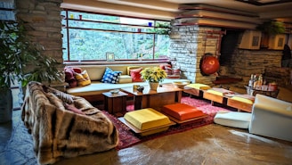 A stylish living room with a mid-century modern aesthetic features stone walls and a large, panoramic window that provides an expansive view of the natural surroundings. The space is furnished with a mix of colorful cushions, low seating, and a central wooden table adorned with a plant. Warm tones and textures, including a plush fur throw, complement the rustic stone fireplace in the background. The atmosphere is cozy and inviting, accentuated by strategically placed decorative items and soft lighting.