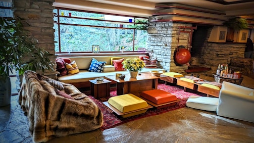 A stylish living room with a mid-century modern aesthetic features stone walls and a large, panoramic window that provides an expansive view of the natural surroundings. The space is furnished with a mix of colorful cushions, low seating, and a central wooden table adorned with a plant. Warm tones and textures, including a plush fur throw, complement the rustic stone fireplace in the background. The atmosphere is cozy and inviting, accentuated by strategically placed decorative items and soft lighting.