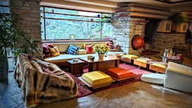 A stylish living room with a mid-century modern aesthetic features stone walls and a large, panoramic window that provides an expansive view of the natural surroundings. The space is furnished with a mix of colorful cushions, low seating, and a central wooden table adorned with a plant. Warm tones and textures, including a plush fur throw, complement the rustic stone fireplace in the background. The atmosphere is cozy and inviting, accentuated by strategically placed decorative items and soft lighting.