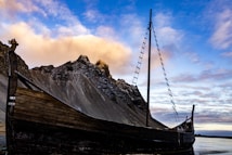 A wooden Viking-style ship is partially grounded on a dark sandy shore, with a mountain range rising dramatically in the background. The sky above is a mix of blue and scattered clouds, with the warm glow of the setting or rising sun casting a golden light.