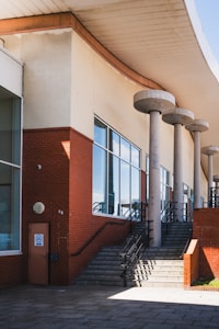 A modern architectural structure featuring large glass windows and red brick walls is depicted. The building has a series of cylindrical concrete columns supporting an overhanging roof. A set of stairs with black railings leads up to a higher level, with sunlight casting shadows across the ground.
