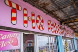 A brightly colored shop front with a large sign featuring red and white candy cane stripes. The words 'The Rock Shop' are prominently displayed against a light pink background. Below the sign, there is a large window and a display promoting candy floss. The area is partially covered by a weathered metal roof, adding a rustic feel.