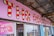A brightly colored shop front with a large sign featuring red and white candy cane stripes. The words 'The Rock Shop' are prominently displayed against a light pink background. Below the sign, there is a large window and a display promoting candy floss. The area is partially covered by a weathered metal roof, adding a rustic feel.
