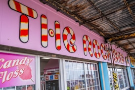A brightly colored shop front with a large sign featuring red and white candy cane stripes. The words 'The Rock Shop' are prominently displayed against a light pink background. Below the sign, there is a large window and a display promoting candy floss. The area is partially covered by a weathered metal roof, adding a rustic feel.