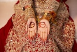 A person wearing an intricate red and gold embellished outfit, adorned with gold bangles and jewelry, displays henna designs on their palms. The henna includes floral patterns and names inscribed within the designs.