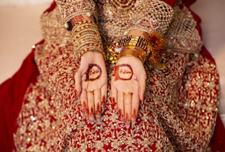 A person wearing an intricate red and gold embellished outfit, adorned with gold bangles and jewelry, displays henna designs on their palms. The henna includes floral patterns and names inscribed within the designs.