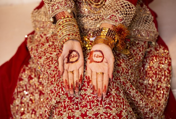 A person wearing an intricate red and gold embellished outfit, adorned with gold bangles and jewelry, displays henna designs on their palms. The henna includes floral patterns and names inscribed within the designs.