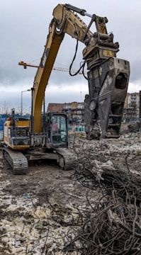 A construction site with a large excavator equipped with a claw attachment is in the foreground. The excavator is working amidst piles of rubble and twisted metal rods in a muddy area. In the background, there are buildings, cranes, and a cloudy sky.