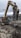 A construction site with a large excavator equipped with a claw attachment is in the foreground. The excavator is working amidst piles of rubble and twisted metal rods in a muddy area. In the background, there are buildings, cranes, and a cloudy sky.