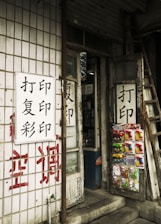 A small shop entrance with worn wooden doors featuring Chinese characters. Various small packaged goods and toys are displayed on one of the doors. The exterior wall is covered with tiles and additional Chinese writing painted in red and black. A ladder leans against the building next to the door.