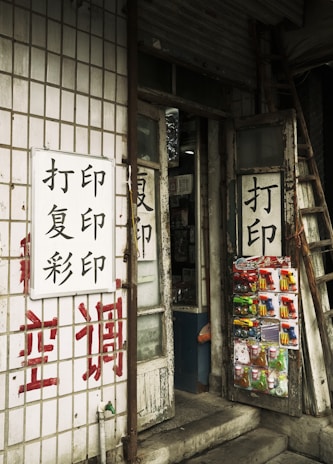 A small shop entrance with worn wooden doors featuring Chinese characters. Various small packaged goods and toys are displayed on one of the doors. The exterior wall is covered with tiles and additional Chinese writing painted in red and black. A ladder leans against the building next to the door.