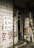 A small shop entrance with worn wooden doors featuring Chinese characters. Various small packaged goods and toys are displayed on one of the doors. The exterior wall is covered with tiles and additional Chinese writing painted in red and black. A ladder leans against the building next to the door.