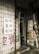 A small shop entrance with worn wooden doors featuring Chinese characters. Various small packaged goods and toys are displayed on one of the doors. The exterior wall is covered with tiles and additional Chinese writing painted in red and black. A ladder leans against the building next to the door.