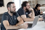 Three people are sitting at a table with laptops, attentively looking at something off camera. The setting appears to be a modern office or classroom, with shelves in the background. The individuals are wearing casual clothing.