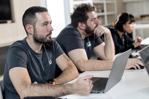 Three people are sitting at a table with laptops, attentively looking at something off camera. The setting appears to be a modern office or classroom, with shelves in the background. The individuals are wearing casual clothing.