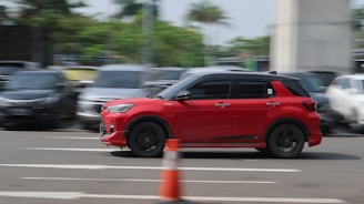 A red car is driving swiftly on a multi-lane road, positioned towards the right side of the image. The background is blurred, indicating motion, with several parked cars and greenery along the sides. A traffic cone is visible in the foreground, suggesting an urban or road setting.
