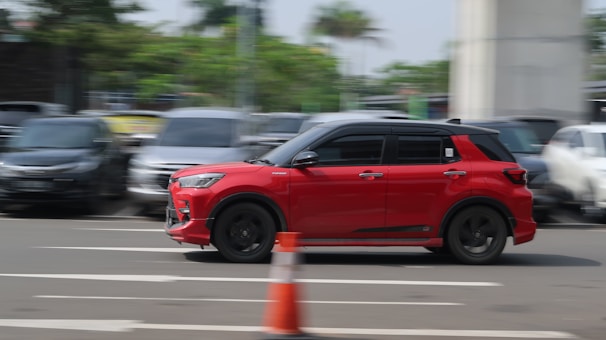 A red car is driving swiftly on a multi-lane road, positioned towards the right side of the image. The background is blurred, indicating motion, with several parked cars and greenery along the sides. A traffic cone is visible in the foreground, suggesting an urban or road setting.
