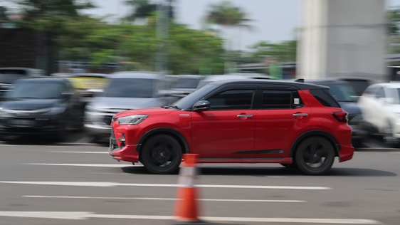 A red car is driving swiftly on a multi-lane road, positioned towards the right side of the image. The background is blurred, indicating motion, with several parked cars and greenery along the sides. A traffic cone is visible in the foreground, suggesting an urban or road setting.