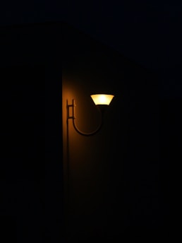 An outdoor wall light with a warm glow illuminates part of a dark building exterior. The light's design is simple and modern, casting a soft yellow hue on the wall.