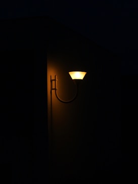 An outdoor wall light with a warm glow illuminates part of a dark building exterior. The light's design is simple and modern, casting a soft yellow hue on the wall.