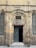 A historic stone building with an arched entranceway displays a sign reading 'Nahhasin School' above the door. The facade features worn brickwork and a blend of stone textures, giving it an ancient appearance. The open wooden door reveals a dark interior. On either side of the entrance, etched plaques are visible with inscriptions.