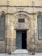 A historic stone building with an arched entranceway displays a sign reading 'Nahhasin School' above the door. The facade features worn brickwork and a blend of stone textures, giving it an ancient appearance. The open wooden door reveals a dark interior. On either side of the entrance, etched plaques are visible with inscriptions.