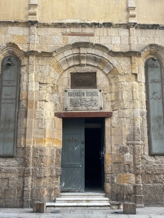 A historic stone building with an arched entranceway displays a sign reading 'Nahhasin School' above the door. The facade features worn brickwork and a blend of stone textures, giving it an ancient appearance. The open wooden door reveals a dark interior. On either side of the entrance, etched plaques are visible with inscriptions.