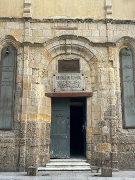 A historic stone building with an arched entranceway displays a sign reading 'Nahhasin School' above the door. The facade features worn brickwork and a blend of stone textures, giving it an ancient appearance. The open wooden door reveals a dark interior. On either side of the entrance, etched plaques are visible with inscriptions.