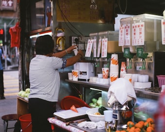 A person in a striped shirt operates a juice vending stall, preparing drinks for customers. The stall features multiple juice dispensers with labels in a foreign language, and a variety of disposable cups stacked next to them. Fresh fruits can be seen on the counter and in baskets, indicating the use of fresh ingredients.