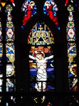 A stained glass window depicting a religious scene featuring a central figure of Jesus Christ on the cross, surrounded by other figures and intricate designs. Angels clad in red are positioned at the top, praying, while other figures are at the sides, looking somber. The background is composed of vibrant, detailed patterns in blue, red, yellow, and green hues.