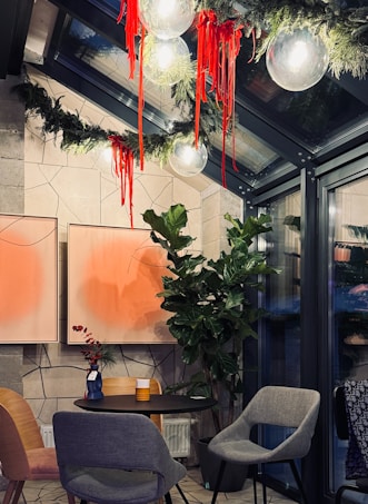 A cozy indoor setting featuring modern decor with two abstract orange paintings on the wall. A small round table is surrounded by stylish chairs, with a potted plant adding greenery to the space. The ceiling is adorned with spherical lights and festive garlands accented by bright red ribbons.
