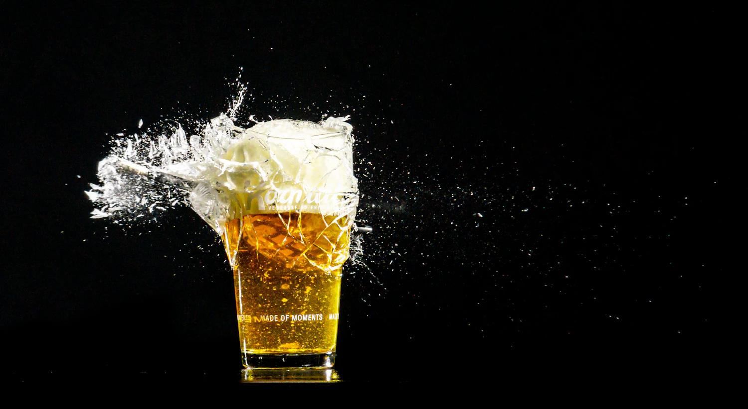 A glass of golden beer exploding with a crown of white foam and spray against a black background