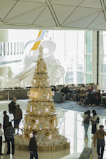 A large decorative Christmas tree stands in the center of a spacious modern indoor area. The tree is adorned with white and gold ornaments including stars and trinkets, and is surrounded by people, some of whom are walking while others are seated in the background. Natural light pours in through large windows, highlighting the metallic and festive elements of the tree.