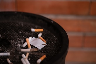 A collection of cigarette butts and ashes discarded in a black ashtray, with a blurred background of reddish-brown bricks.