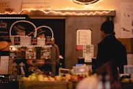 A cozy market stall featuring a variety of items such as jars of preserves and baked goods is displayed. Two individuals are present, one behind a counter working and another customer facing them. The stall is decorated with warm fairy lights, adding an inviting atmosphere. Signs in Italian and promotional materials are visible, with fruits and other goods neatly arranged in the foreground.