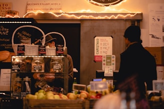 A cozy market stall featuring a variety of items such as jars of preserves and baked goods is displayed. Two individuals are present, one behind a counter working and another customer facing them. The stall is decorated with warm fairy lights, adding an inviting atmosphere. Signs in Italian and promotional materials are visible, with fruits and other goods neatly arranged in the foreground.