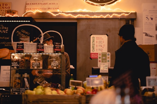A cozy market stall featuring a variety of items such as jars of preserves and baked goods is displayed. Two individuals are present, one behind a counter working and another customer facing them. The stall is decorated with warm fairy lights, adding an inviting atmosphere. Signs in Italian and promotional materials are visible, with fruits and other goods neatly arranged in the foreground.