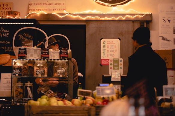 A cozy market stall featuring a variety of items such as jars of preserves and baked goods is displayed. Two individuals are present, one behind a counter working and another customer facing them. The stall is decorated with warm fairy lights, adding an inviting atmosphere. Signs in Italian and promotional materials are visible, with fruits and other goods neatly arranged in the foreground.